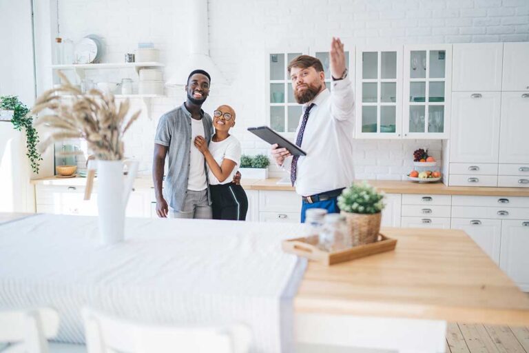 A real estate agent reviews paperwork with a couple at a kitchen table, with documents and a laptop visible.