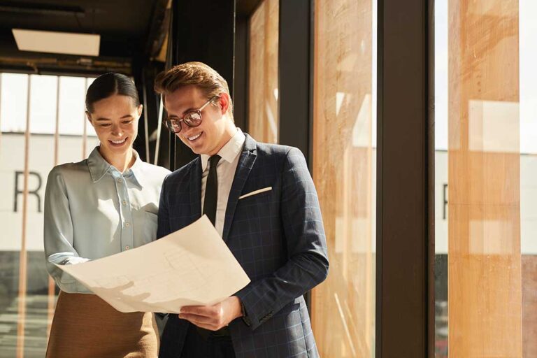 A couple sits at a kitchen table asking questions to a real estate agent with folders and paperwork.